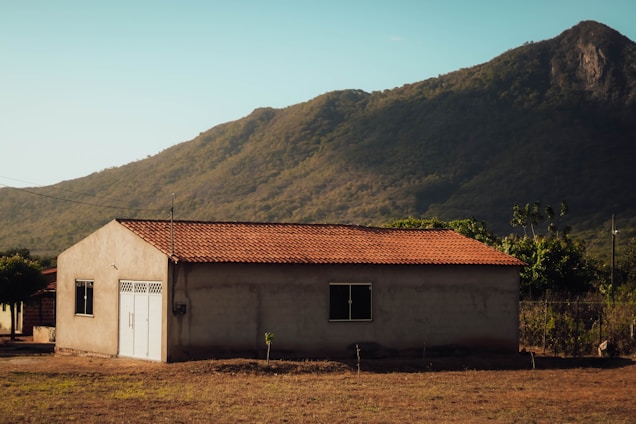 A solitary house with a reddish-brown tiled roof stands in a serene rural setting. It is surrounded by sparse vegetation and open land, with a backdrop of green, forested hills under a clear blue sky.