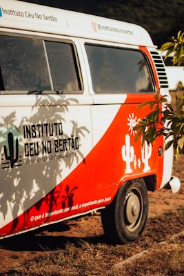 A vintage van painted in white and red with cactus and sun designs on the side. The text 'Instituto Céu No Sertão' is visible, along with Instagram handle details and a message in Portuguese. The van is located outdoors with foliage partially shading it.
