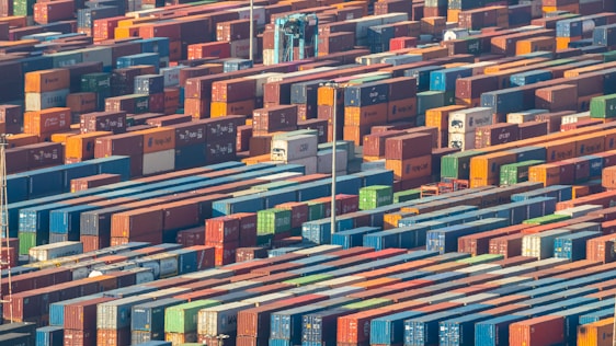 Numerous shipping containers are stacked in an organized manner, filling the entire frame. They vary in colors, such as red, blue, green, and orange, creating a vibrant and busy atmosphere. A crane is seen among the containers, indicating an industrial setting.