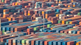 Numerous shipping containers are stacked in an organized manner, filling the entire frame. They vary in colors, such as red, blue, green, and orange, creating a vibrant and busy atmosphere. A crane is seen among the containers, indicating an industrial setting.