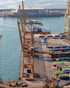 Overhead view of a busy port featuring large yellow cranes standing alongside a dock. Stacked colorful shipping containers in various hues like green, blue, and red are neatly arranged, indicating an active shipping operation. Calm water reflects the light, and a distant terminal building with a modern design is visible, suggesting an infrastructural hub.