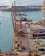 Overhead view of a busy port featuring large yellow cranes standing alongside a dock. Stacked colorful shipping containers in various hues like green, blue, and red are neatly arranged, indicating an active shipping operation. Calm water reflects the light, and a distant terminal building with a modern design is visible, suggesting an infrastructural hub.