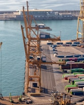 Overhead view of a busy port featuring large yellow cranes standing alongside a dock. Stacked colorful shipping containers in various hues like green, blue, and red are neatly arranged, indicating an active shipping operation. Calm water reflects the light, and a distant terminal building with a modern design is visible, suggesting an infrastructural hub.