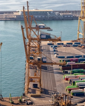 Overhead view of a busy port featuring large yellow cranes standing alongside a dock. Stacked colorful shipping containers in various hues like green, blue, and red are neatly arranged, indicating an active shipping operation. Calm water reflects the light, and a distant terminal building with a modern design is visible, suggesting an infrastructural hub.