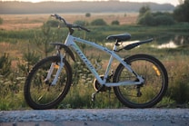 A white mountain bike with black wheels stands on a gravel path in a lush, green countryside setting. The background showcases an expansive view of fields, trees, and a serene body of water under a clear sky, subtly lit by the soft glow of a setting or rising sun.
