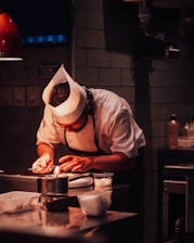 A chef wearing a white hat and uniform is deeply focused on using a small torch to prepare a dish in a dimly lit kitchen. Various kitchen utensils and ingredients are visible on the countertop.