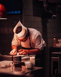 A chef wearing a white hat and uniform is deeply focused on using a small torch to prepare a dish in a dimly lit kitchen. Various kitchen utensils and ingredients are visible on the countertop.