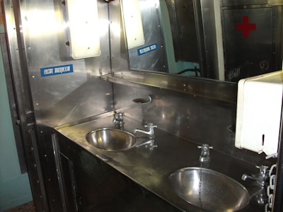 A metal washroom interior featuring two stainless steel sinks with attached faucets and chains. The sinks are built into a countertop against the wall, which has a mirror and a sign labeled 'CREWS WASHROOM'. A paper towel dispenser is visible to the right, and there is a red cross symbol on a cabinet in the background.