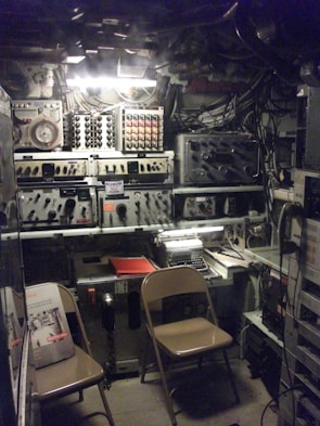 A dimly lit room filled with vintage radio and communication equipment. There are various control panels with dials and switches, a typewriter on a desk, and several wires and cables visible. Two folding chairs are positioned in front of the equipment, and manuals or booklets are placed on one of the chairs.