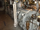 A well-maintained industrial or mechanical room featuring a variety of metal machinery and pipes. There is a large valve wheel and various gauges and switches, suggesting it may be part of a ship's engine room or an industrial plant.