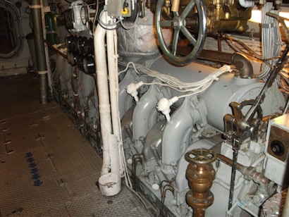 A well-maintained industrial or mechanical room featuring a variety of metal machinery and pipes. There is a large valve wheel and various gauges and switches, suggesting it may be part of a ship's engine room or an industrial plant.