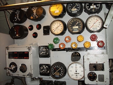 A collection of analog gauges, dials, and switches mounted on a metal panel. The gauges display various measurements such as pressure, voltage, and RPM, with colored knobs and switches below them, indicating control functionality. The control panel shows signs of wear, suggesting frequent use.
