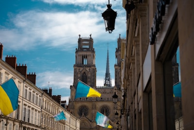 A picturesque urban street view featuring a large cathedral with twin towers and a spire in the background. The street is lined with old buildings with Ukrainian flags hanging outside them. There are several street lamps attached to the buildings, and the sky is partly cloudy.