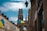 A picturesque urban street view featuring a large cathedral with twin towers and a spire in the background. The street is lined with old buildings with Ukrainian flags hanging outside them. There are several street lamps attached to the buildings, and the sky is partly cloudy.