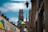 A picturesque urban street view featuring a large cathedral with twin towers and a spire in the background. The street is lined with old buildings with Ukrainian flags hanging outside them. There are several street lamps attached to the buildings, and the sky is partly cloudy.