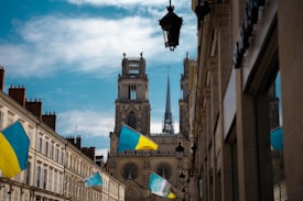 A picturesque urban street view featuring a large cathedral with twin towers and a spire in the background. The street is lined with old buildings with Ukrainian flags hanging outside them. There are several street lamps attached to the buildings, and the sky is partly cloudy.