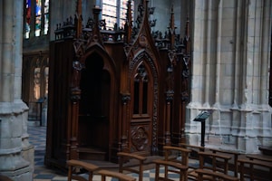 Intricately carved wooden confessional booth set in a stone-walled church interior, featuring pointed arches and stained glass windows in the background. Wooden pews are lined up in the foreground, enhancing the solemn and sacred atmosphere.