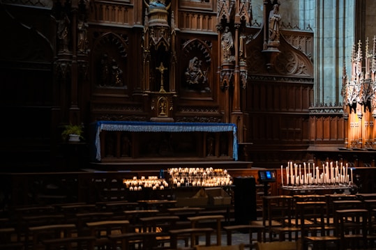 The image depicts the interior of a church with an ornately carved wooden altar as the focal point. Candles are lit in front of the altar, creating a warm, inviting glow in the otherwise dimly lit space. Wooden pews are arranged in rows, and intricate architectural details adorn the surrounding walls.