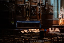 The image depicts the interior of a church with an ornately carved wooden altar as the focal point. Candles are lit in front of the altar, creating a warm, inviting glow in the otherwise dimly lit space. Wooden pews are arranged in rows, and intricate architectural details adorn the surrounding walls.