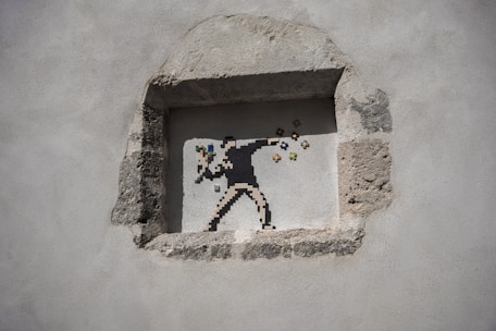 A pixel art mural depicting a figure in a throwing stance, reminiscent of a protester, but instead of a projectile, colorful flowers emerge from the hand. The artwork is situated within a rough-hewn stone niche on a plain wall.