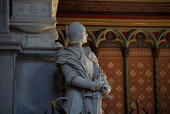 A detailed marble statue of a knight in armor is positioned against an ornate, decorative wall. The knight is holding a sword and looking upwards, capturing a sense of contemplation or vigilance. The background features intricate patterns and arches in warm, muted colors.