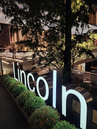 A luminous sign with the word 'Lincoln' stands prominently amid a lush setting of bushes and trees. Behind the foliage, a modern urban structure with glowing horizontal lights creates a sophisticated backdrop. The warm light contrasts with the natural greenery.