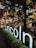A luminous sign with the word 'Lincoln' stands prominently amid a lush setting of bushes and trees. Behind the foliage, a modern urban structure with glowing horizontal lights creates a sophisticated backdrop. The warm light contrasts with the natural greenery.