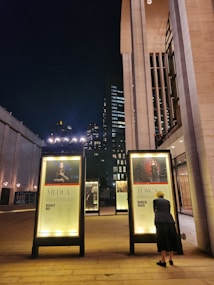 A nighttime urban scene featuring a modern architectural structure with illuminated posters promoting opera performances of Medea and Tosca. A person is closely looking at one of the posters. The background includes tall buildings and city lights.