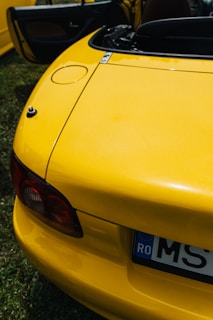 A close-up of the rear section of a bright yellow convertible car with its trunk slightly open. The car has a European style license plate with visible characters, and part of the interior is visible through the open door. The photo is taken in daylight and the car is parked on grass.