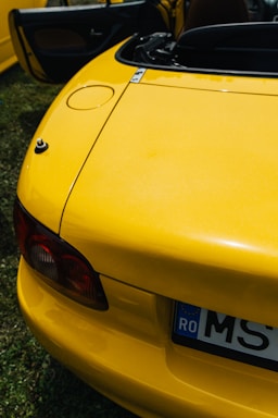 A close-up of the rear section of a bright yellow convertible car with its trunk slightly open. The car has a European style license plate with visible characters, and part of the interior is visible through the open door. The photo is taken in daylight and the car is parked on grass.