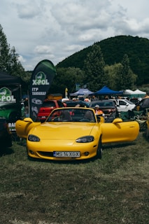 A bright yellow convertible sports car is parked on grass, surrounded by several other vehicles and tents. People are interacting in the background, suggesting a car show or outdoor event. There are banners and flags displaying the X-POL logo.
