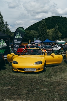 A bright yellow convertible sports car is parked on grass, surrounded by several other vehicles and tents. People are interacting in the background, suggesting a car show or outdoor event. There are banners and flags displaying the X-POL logo.