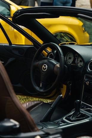 An interior view of a convertible sports car, featuring a detailed dashboard with three round gauges and a steering wheel adorned with the Mazda logo. The gear stick is visible, and the interior is predominantly black with brown seating. Another yellow car is visible in the background.