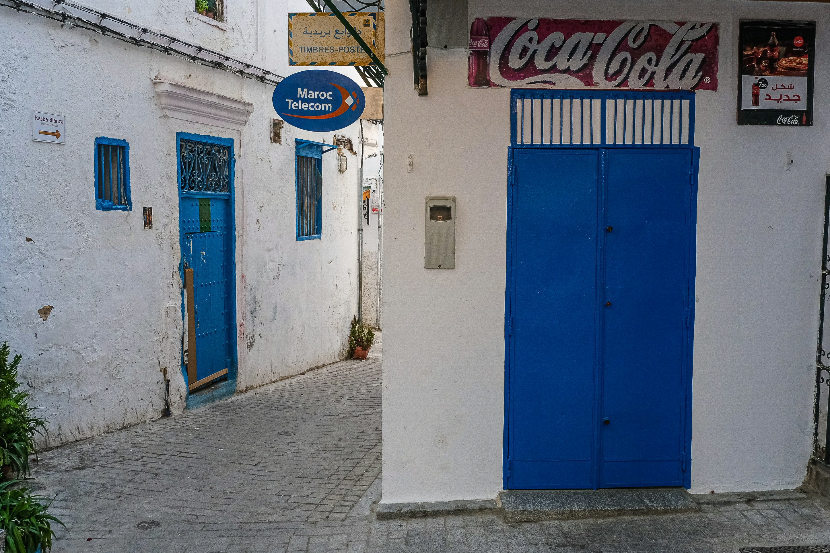 a blue and white building with a blue door, A narrow street in Tangier, Morocco. The scene features whitewashed buildings with vibrant blue doors and window frames, typical of Moroccan architecture. Signs for Maroc Telecom and Coca-Cola add a touch of modernity to the traditional setting. The alleyway is paved with cobblestones, leading around a corner and inviting exploration.