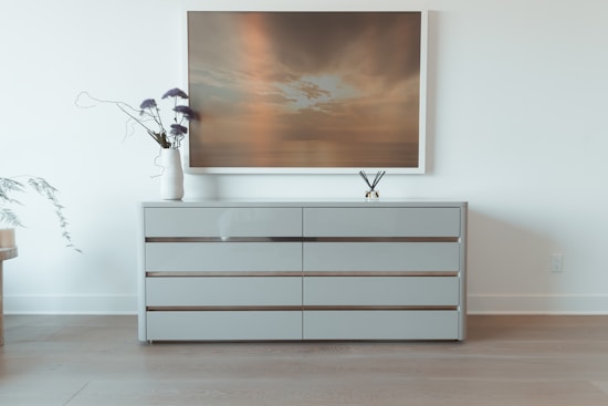 A sleek, minimalist dresser with three horizontal drawers sits against a light-colored wall. Above the dresser hangs a large framed artwork depicting a tranquil sunset over water. On the dresser, there is a white vase with purple flowers and a small diffuser with reed sticks.