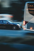 A motion blur effect captures a black car speeding past a large white bus on a city street. The background appears dark, with lights from other vehicles creating streaks, conveying a sense of movement and urgency.