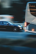A motion blur effect captures a black car speeding past a large white bus on a city street. The background appears dark, with lights from other vehicles creating streaks, conveying a sense of movement and urgency.