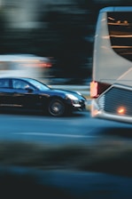 A motion blur effect captures a black car speeding past a large white bus on a city street. The background appears dark, with lights from other vehicles creating streaks, conveying a sense of movement and urgency.