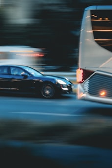 A motion blur effect captures a black car speeding past a large white bus on a city street. The background appears dark, with lights from other vehicles creating streaks, conveying a sense of movement and urgency.