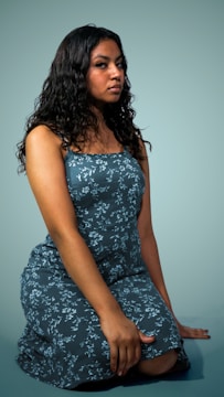 A person with long, wavy, dark hair is posing, wearing a sleeveless dress with a floral pattern. The dress is predominantly blue, and the person is kneeling on one knee against a simple, solid background.