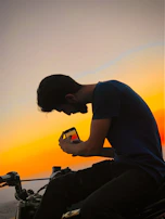 Sunset silhouette of a biker standing beside a motorcycle, holding a leather bag.