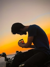 A friendly biker holding a phone, smiling warmly in front of a motorcycle with a sunset background.