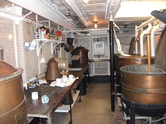 A room filled with various copper kettles and machinery, likely used for brewing or distilling. Shelves are lined with jars and containers, while white mugs hang from hooks. The background features mesh fencing and pipes along the ceiling.
