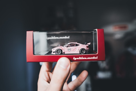 A hand is holding a boxed model car with a pink color and racing decals. The model car is enclosed in a display case with a red border featuring the text 'ignition.model.' The background is blurred, drawing focus to the detailed toy car.