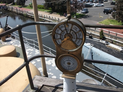 A ship's engine telegraph is mounted prominently on a deck, offering commands like full, half, and stop. The scene includes a waterway and marina with cars parked in the distance, along with some green areas and trees.