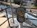 A ship's engine telegraph is mounted prominently on a deck, offering commands like full, half, and stop. The scene includes a waterway and marina with cars parked in the distance, along with some green areas and trees.