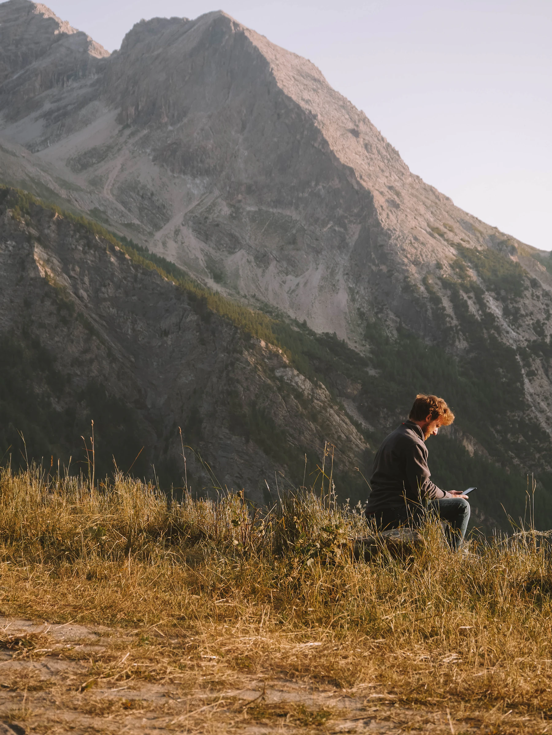A man sitting reading on a grassy ridge below a mountain