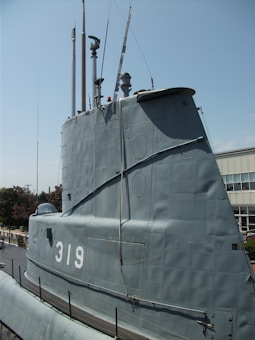 A large, metallic submarine structure with the number 319 prominently displayed. Several vertical poles and antennas protrude from the top, indicative of a naval vessel docked at a port. The background shows a clear sky and a building, suggesting the submarine is on display or stationed near an urban location.