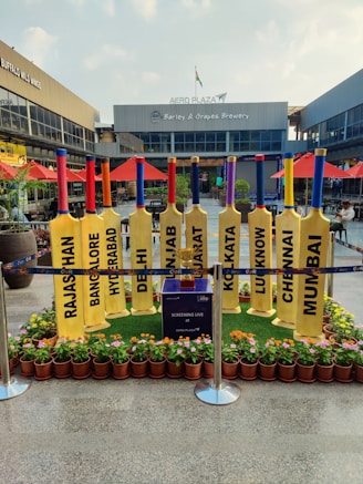 A row of large, yellow cricket bats is displayed outdoors, each with the name of an Indian city printed on it. The setting is a shopping plaza with storefronts like Buffalo Wild Wings and Barley & Grapes Brewery visible in the background. Red umbrellas and small potted plants decorate the area. The space is cordoned off with blue barriers, and a small table or box in the center mentions a live screening at Aero Plaza.