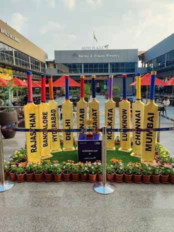 A row of large, yellow cricket bats is displayed outdoors, each with the name of an Indian city printed on it. The setting is a shopping plaza with storefronts like Buffalo Wild Wings and Barley & Grapes Brewery visible in the background. Red umbrellas and small potted plants decorate the area. The space is cordoned off with blue barriers, and a small table or box in the center mentions a live screening at Aero Plaza.